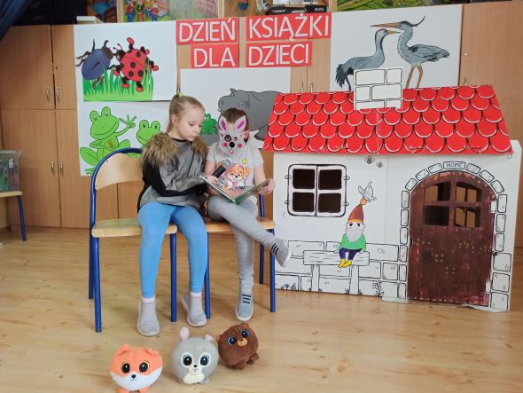  Zdjęcie przedstawia dzieci siedzące i czytające książki, w tle domek, rysunki i hasło Dzień Ksiązki dla dzieci