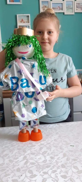 grafika przedstawia dziewczynkę z wykonaną lalką. Lalka ma na głowie ma burze loków w kolorze zielonym. Ubrana jest w sukienkę z plastikowej foli z nadrukiem, przepasana jest torebką wykonaną z wstążki i kubeczków po serku. Buciki lalki to pomarańczowe nakrętki.
